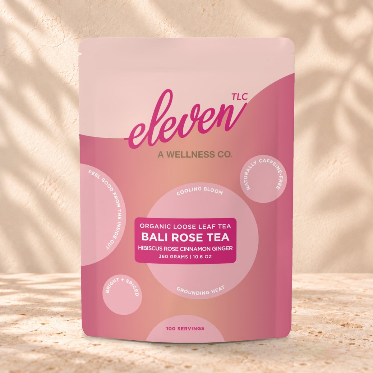 Eleven TLC Bali Rose Tea 360g pink and coral pouch standing on a travertine ledge against warm plaster, with a contact shadow beneath the base and a soft shadow to the right.