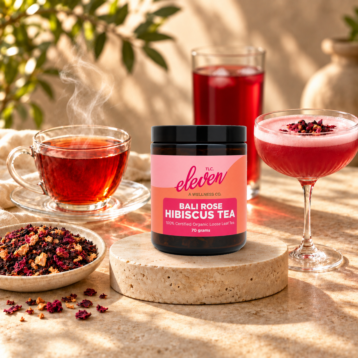 Eleven TLC Bali Rose Hibiscus Tea 70g jar on a travertine riser beside hot tea, iced ruby tea, a pink botanical mocktail and loose-leaf tea.