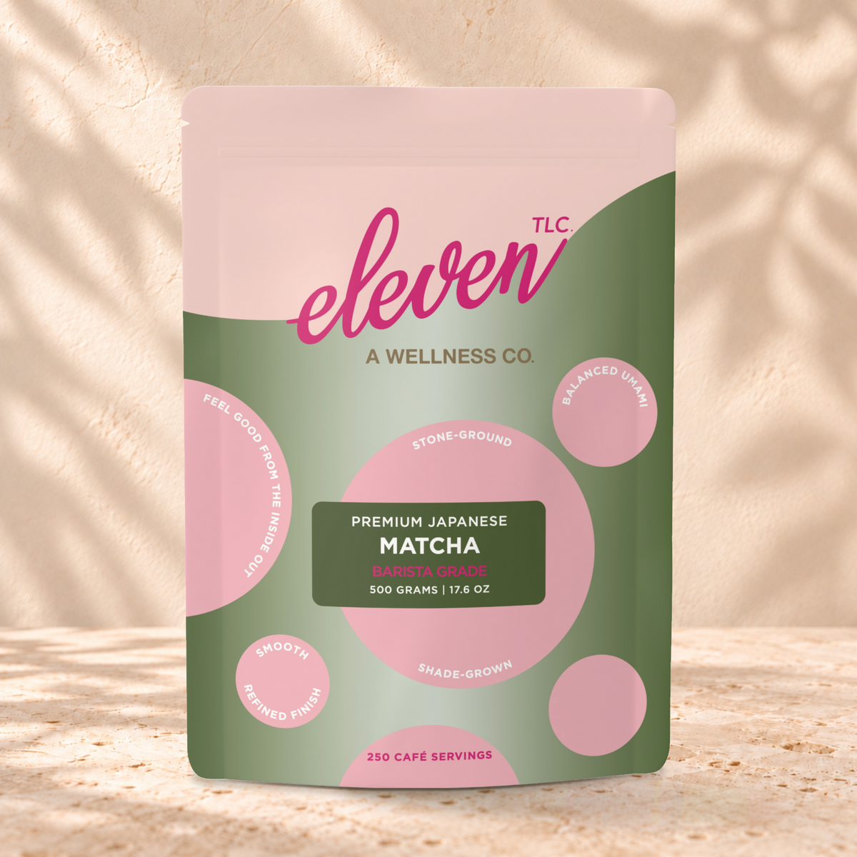 Eleven TLC Barista Matcha 500g green and pink pouch standing on a travertine ledge against warm plaster, with a contact shadow beneath the base and a soft shadow to the right.