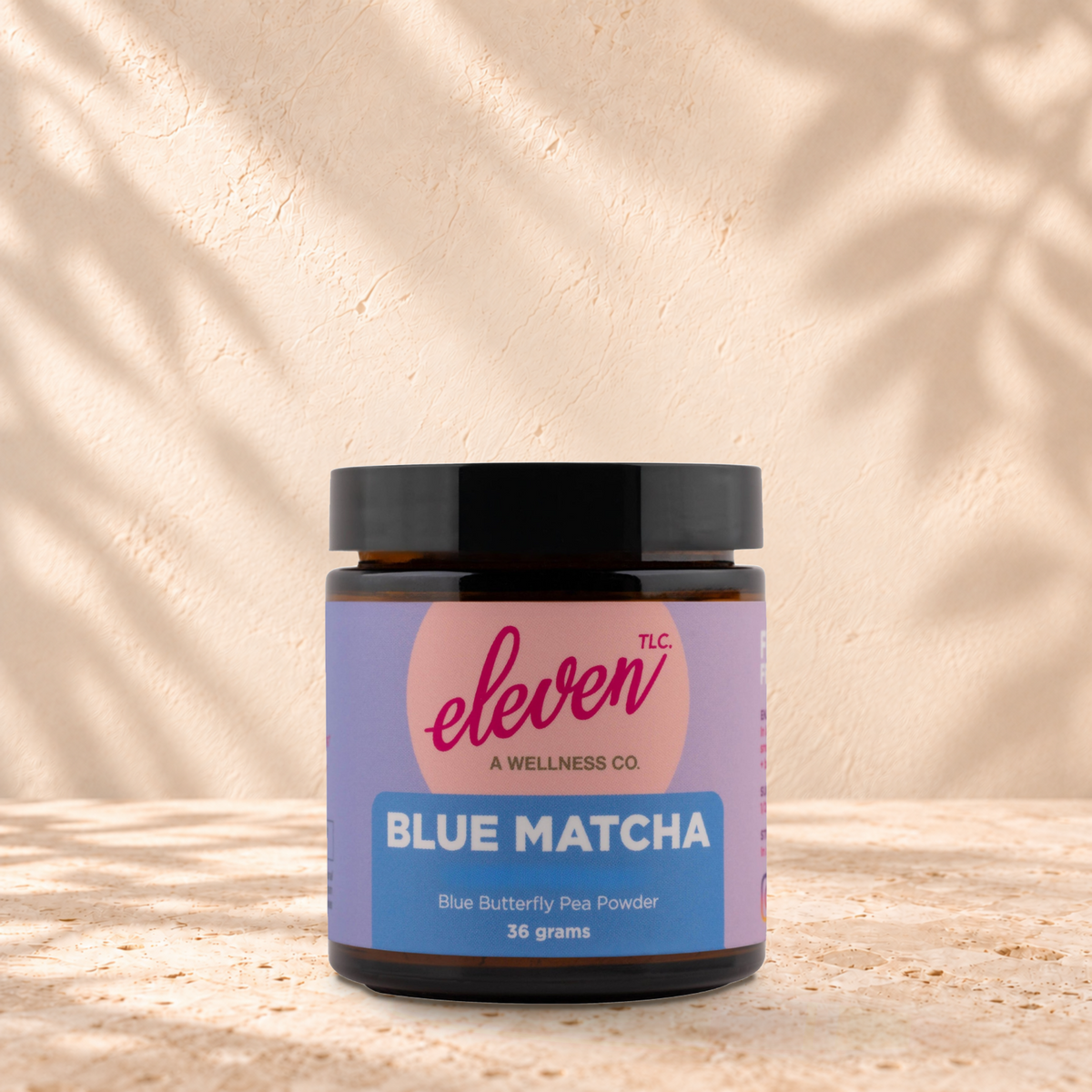 Eleven TLC Blue Matcha butterfly pea flower powder 36g amber jar with a black lid standing on a travertine ledge against warm plaster, with a contact shadow beneath its base and a soft shadow to the right.