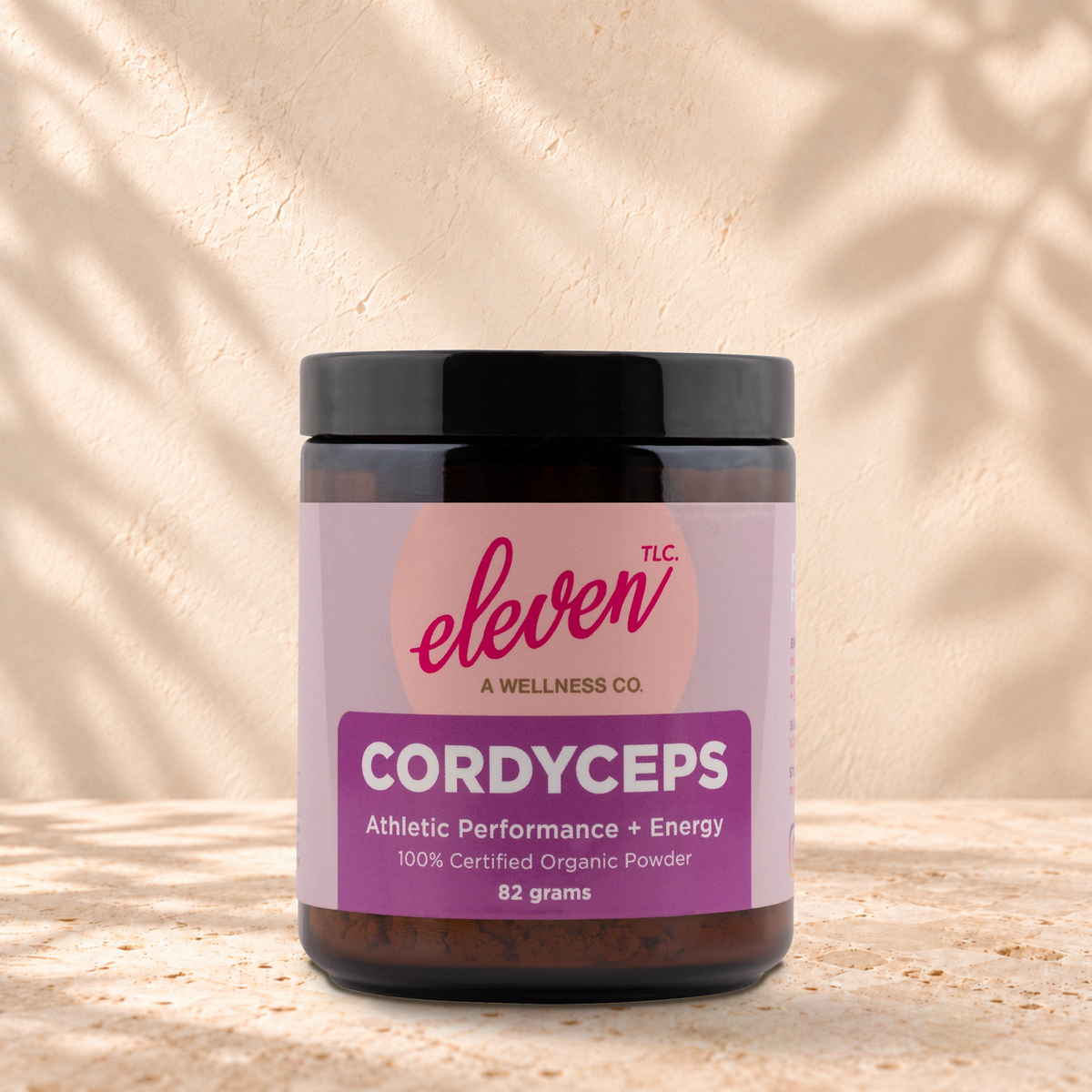 Eleven TLC Organic Cordyceps Mushroom Powder | Full-Spectrum, 82g jar in an amber glass jar with a black lid on a travertine ledge against warm plaster, with a slightly stronger contact shadow beneath the base and a soft shadow to the right.