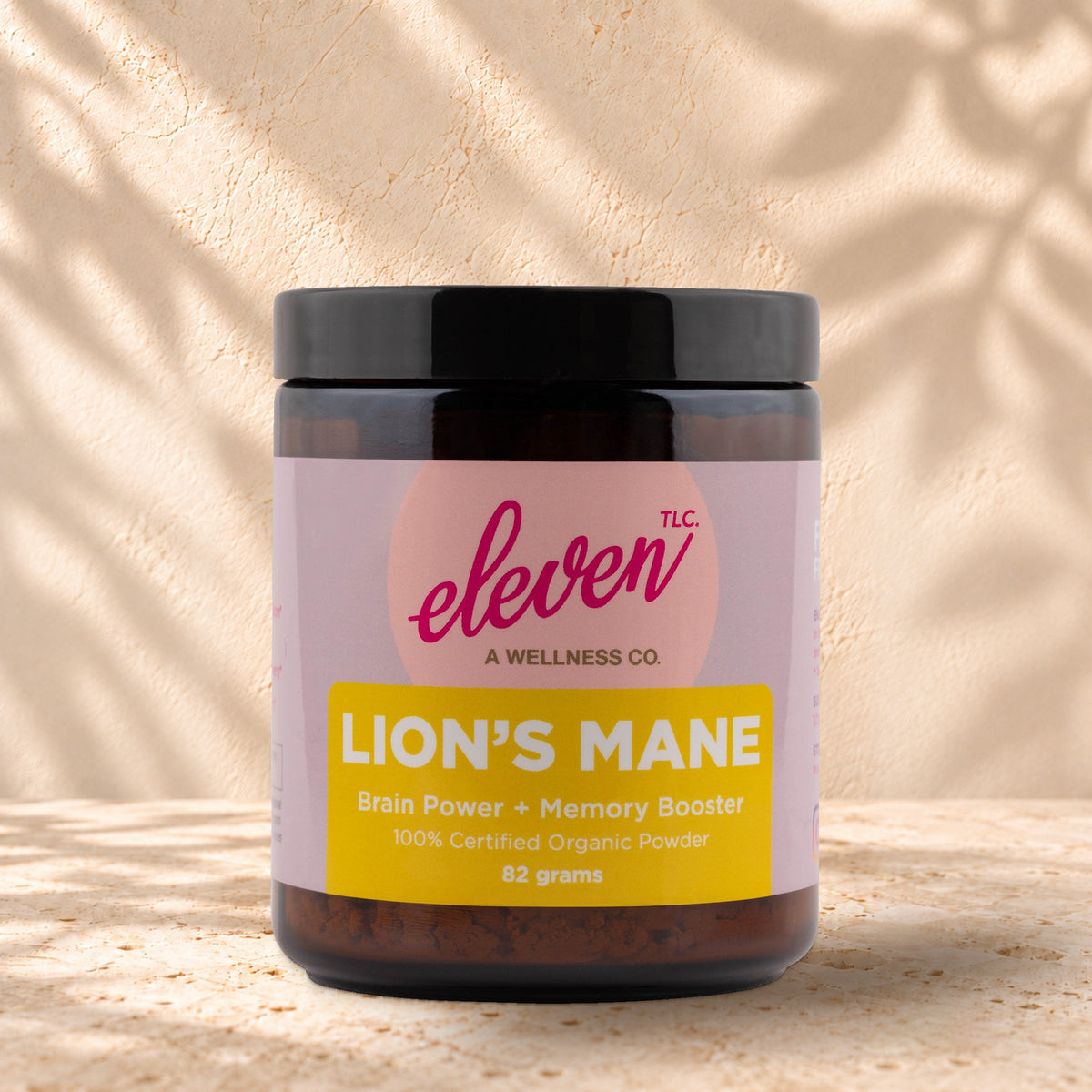 Front view of an Eleven TLC Lion’s Mane 82g amber jar with a black lid on warm travertine against a cream plaster wall, with a soft shadow extending right.