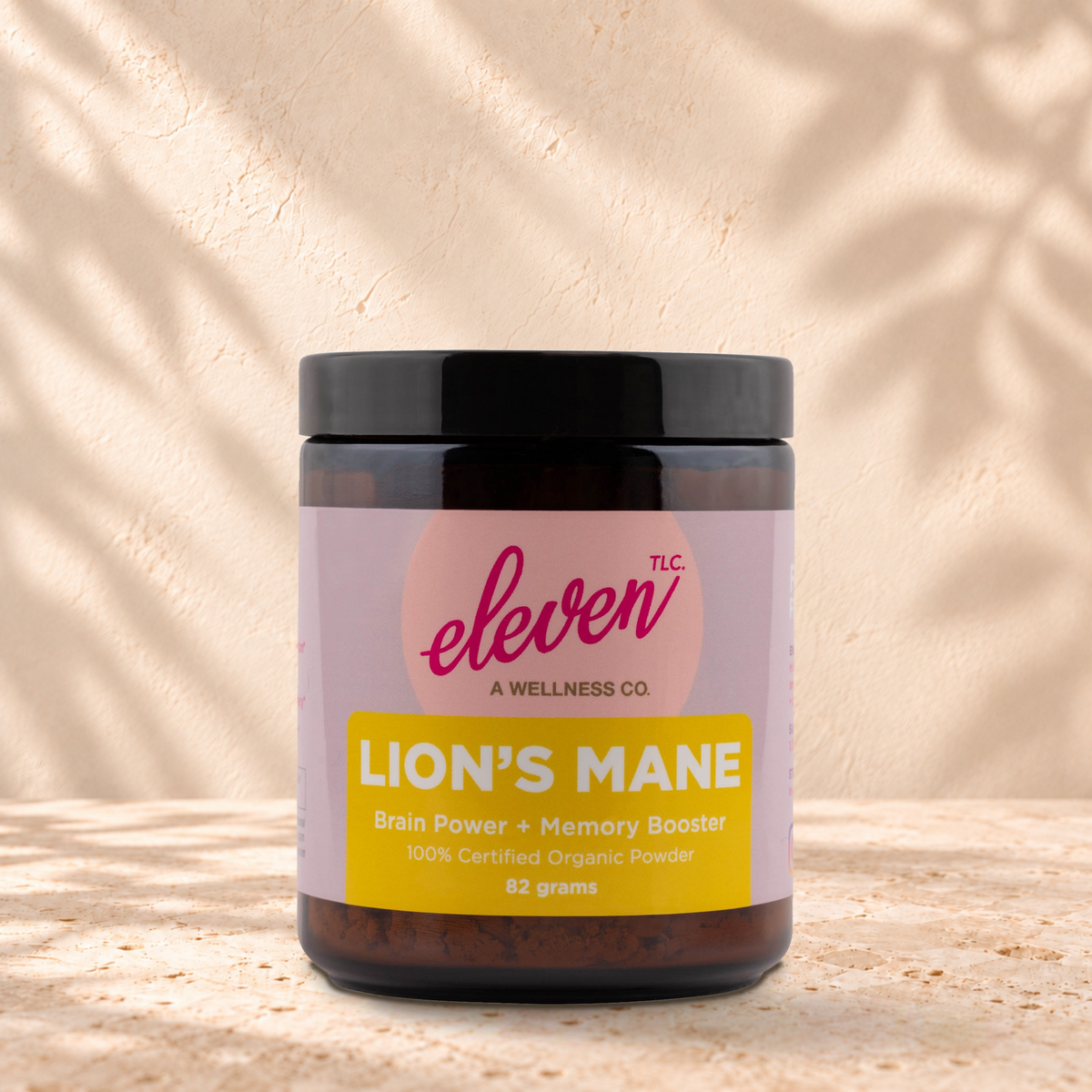 Eleven TLC Organic Lion's Mane Mushroom Powder | Focus & Clarity, 82g jar in an amber glass jar with a black lid on a travertine ledge against warm plaster, with a slightly stronger contact shadow beneath the base and a soft shadow to the right.