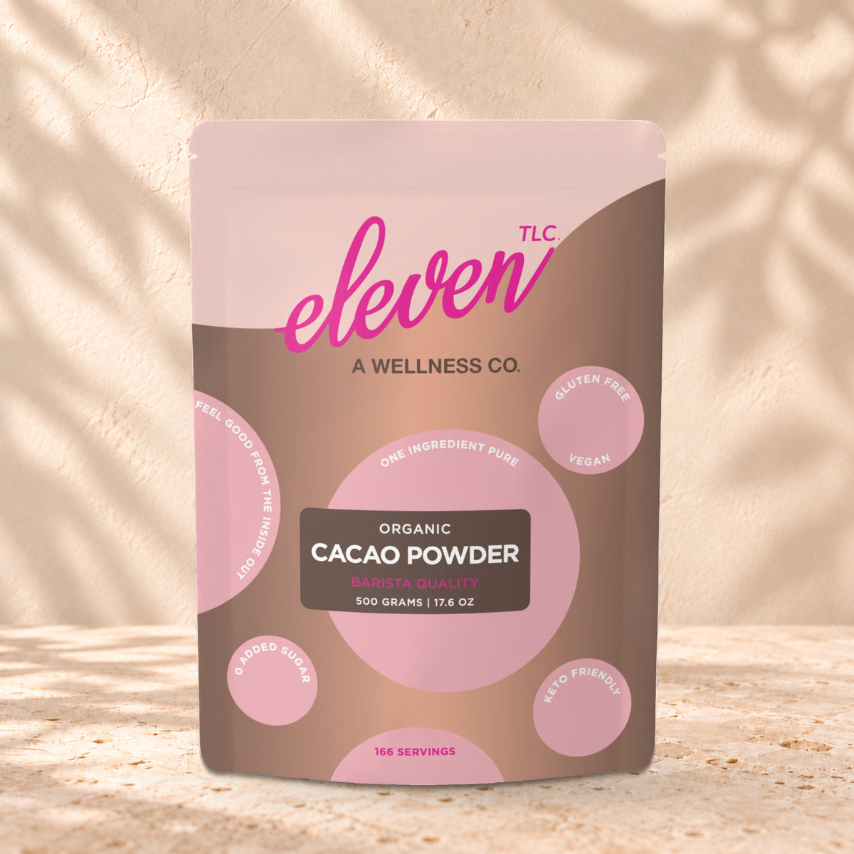 Eleven TLC Organic Cacao Powder 500g stand-up pouch on travertine stone against a warm cream plaster wall, lit from the left with a soft shadow to the right.