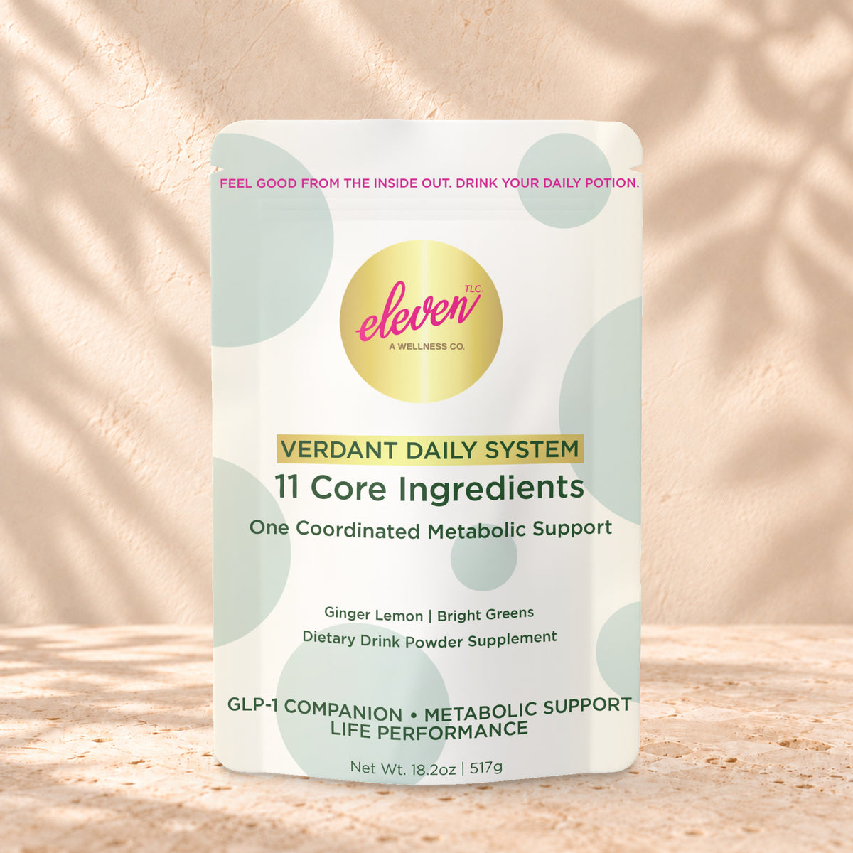 Eleven TLC Verdant Daily Metabolic System 517g stand-up pouch on travertine stone against a warm cream plaster wall, lit from the left with a soft shadow to the right.