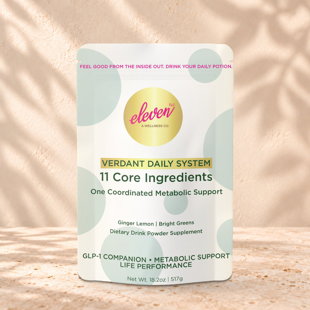 Eleven TLC Verdant Daily Metabolic System 517g stand-up pouch on travertine stone against a warm cream plaster wall, lit from the left with a soft shadow to the right.