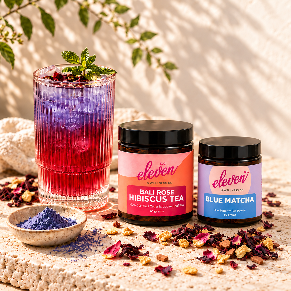 Bali Bloom iced hibiscus tea with violet butterfly pea ice beside Bali Rose and Blue Matcha jars on cream travertine.