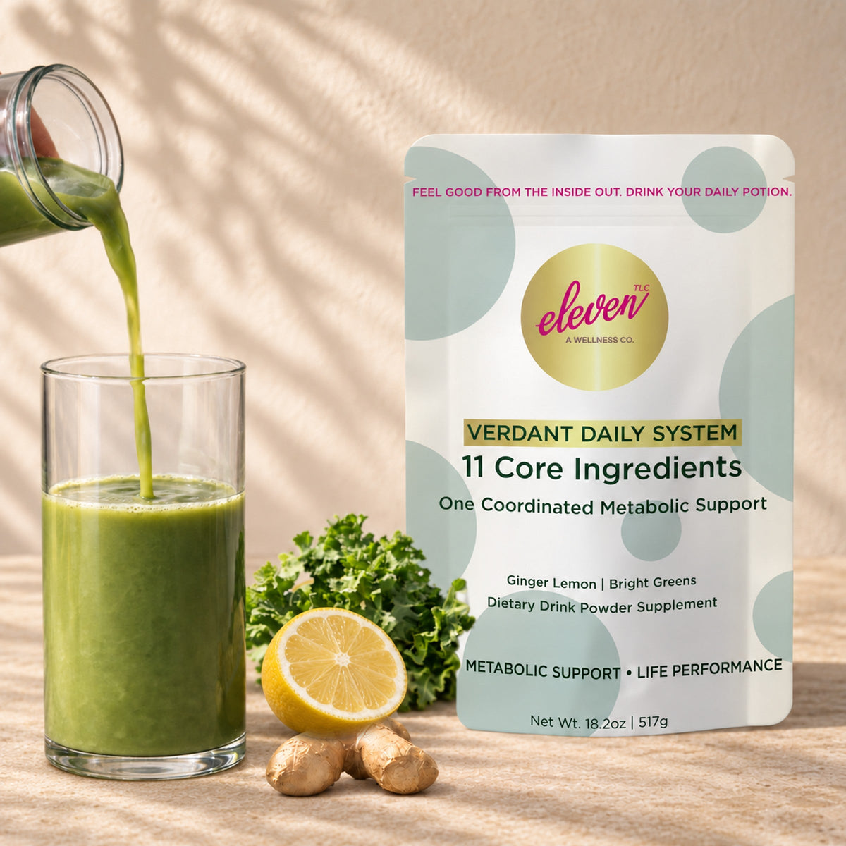 Green drink being poured into a glass beside an Eleven TLC Verdant Daily System pouch, lemon, ginger, and kale on sunlit stone.