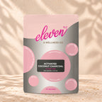 Eleven TLC Activated Coconut Charcoal Powder 500g stand-up pouch on travertine stone against a warm cream plaster wall, lit from the left with a soft shadow to the right.