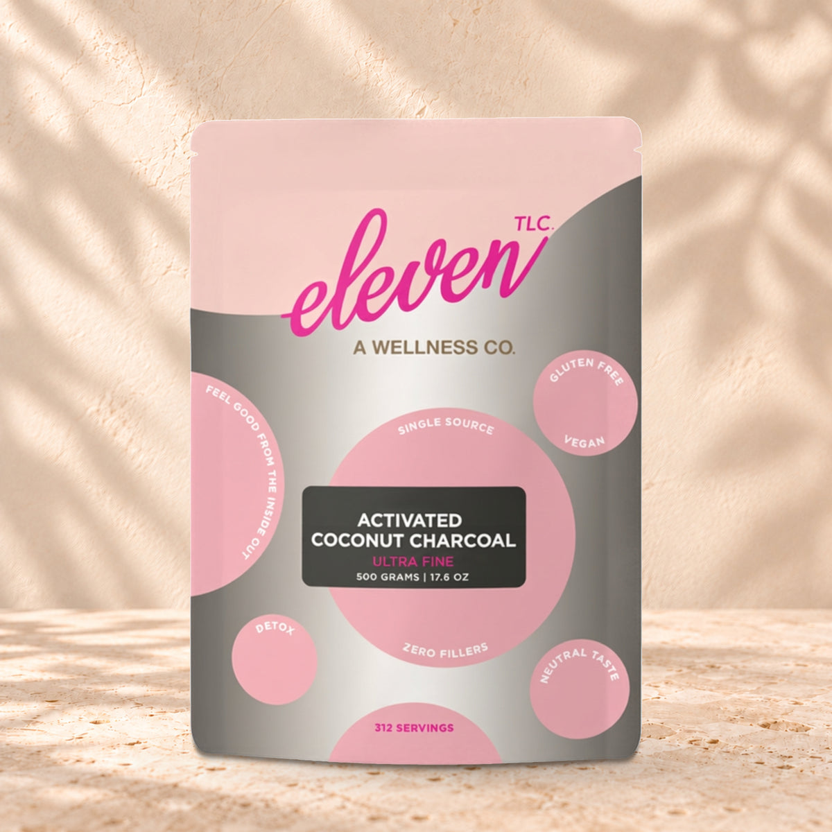 Eleven TLC Activated Coconut Charcoal Powder 500g stand-up pouch on travertine stone against a warm cream plaster wall, lit from the left with a soft shadow to the right.