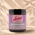 Front view of an Eleven TLC Ashwagandha 120g amber jar with a black lid on warm travertine against a cream plaster wall, with a soft shadow extending right.