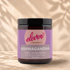 Front view of an Eleven TLC Ashwagandha 120g amber jar with a black lid on warm travertine against a cream plaster wall, with a soft shadow extending right.