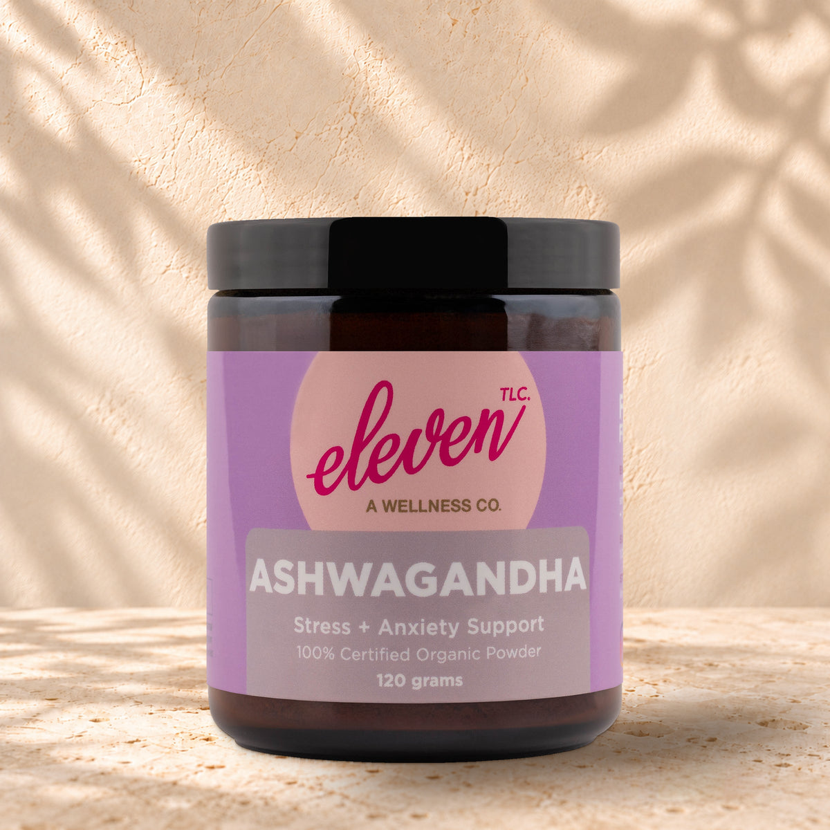 Front view of an Eleven TLC Ashwagandha 120g amber jar with a black lid on warm travertine against a cream plaster wall, with a soft shadow extending right.