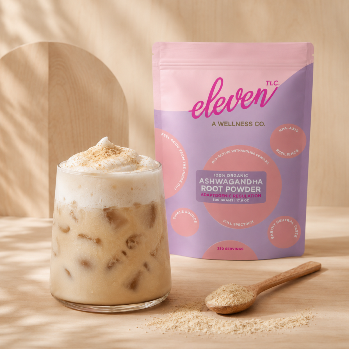 Eleven TLC Ashwagandha Root Powder 500g pouch beside a frothy iced latte and a wooden spoon of powder on a warm wood surface.
