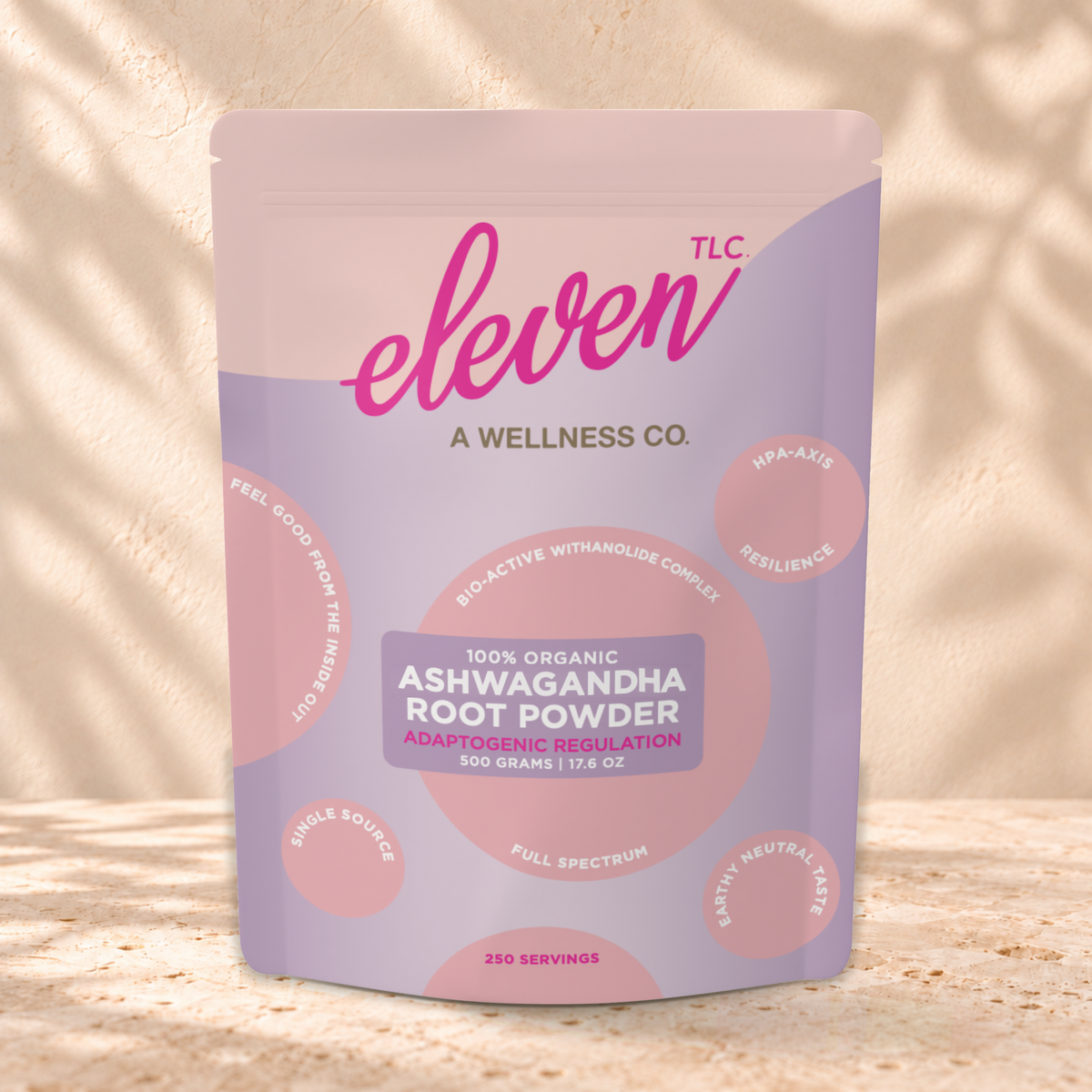 Eleven TLC Ashwagandha Root Powder 500g lavender and pink pouch standing on a travertine ledge against warm plaster, with a contact shadow beneath the base and a soft shadow to the right.