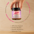 Hand lifting the Eleven TLC ashwagandha root powder 120g amber glass jar, ringed by the words gluten free, vegan, non-GMO certified, certified organic, full spectrum and 3rd party lab tested.