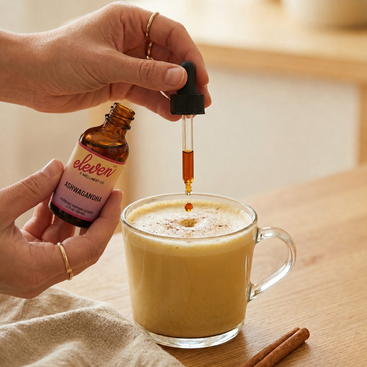 Hand dispensing ashwagandha liquid extract into a ginger turmeric latte in a clear glass mug on a wooden surface.