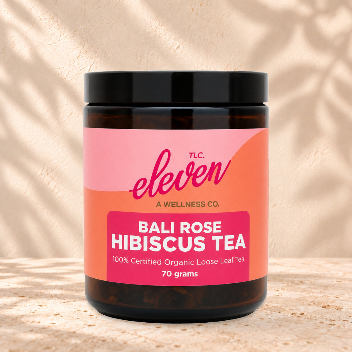 Eleven TLC Bali Rose Loose Leaf Tea 70g amber glass jar with a black lid on travertine stone against a warm cream plaster wall, with a soft shadow to the right.