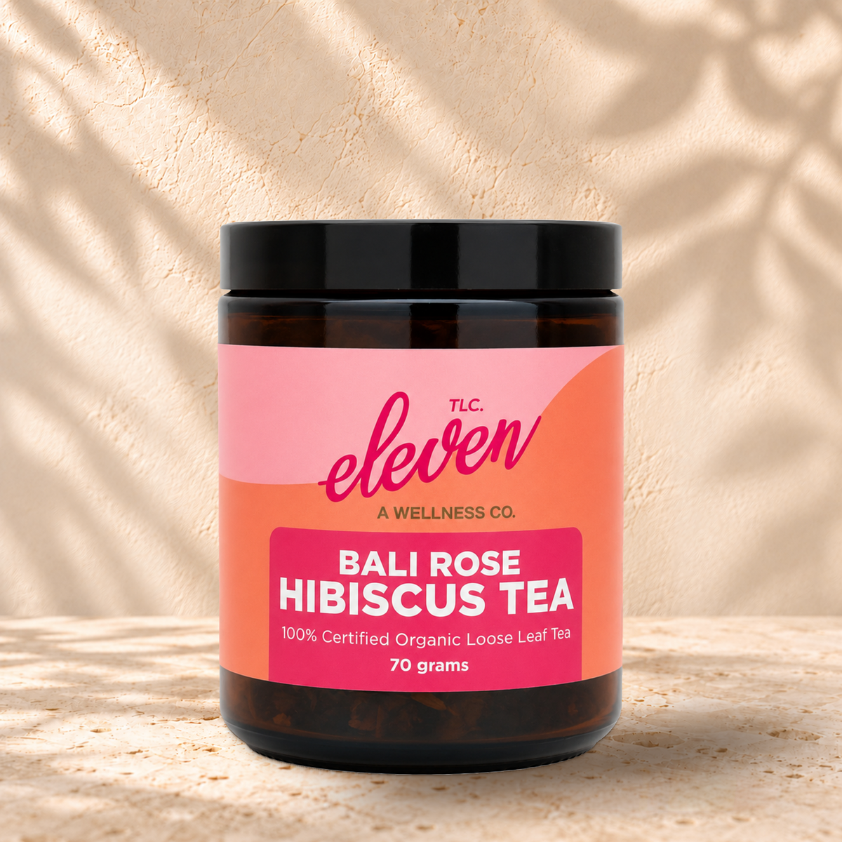 Front view of an Eleven TLC Bali Rose Hibiscus Tea 70g amber jar with a black lid and pink and coral label on warm travertine against a cream plaster wall.