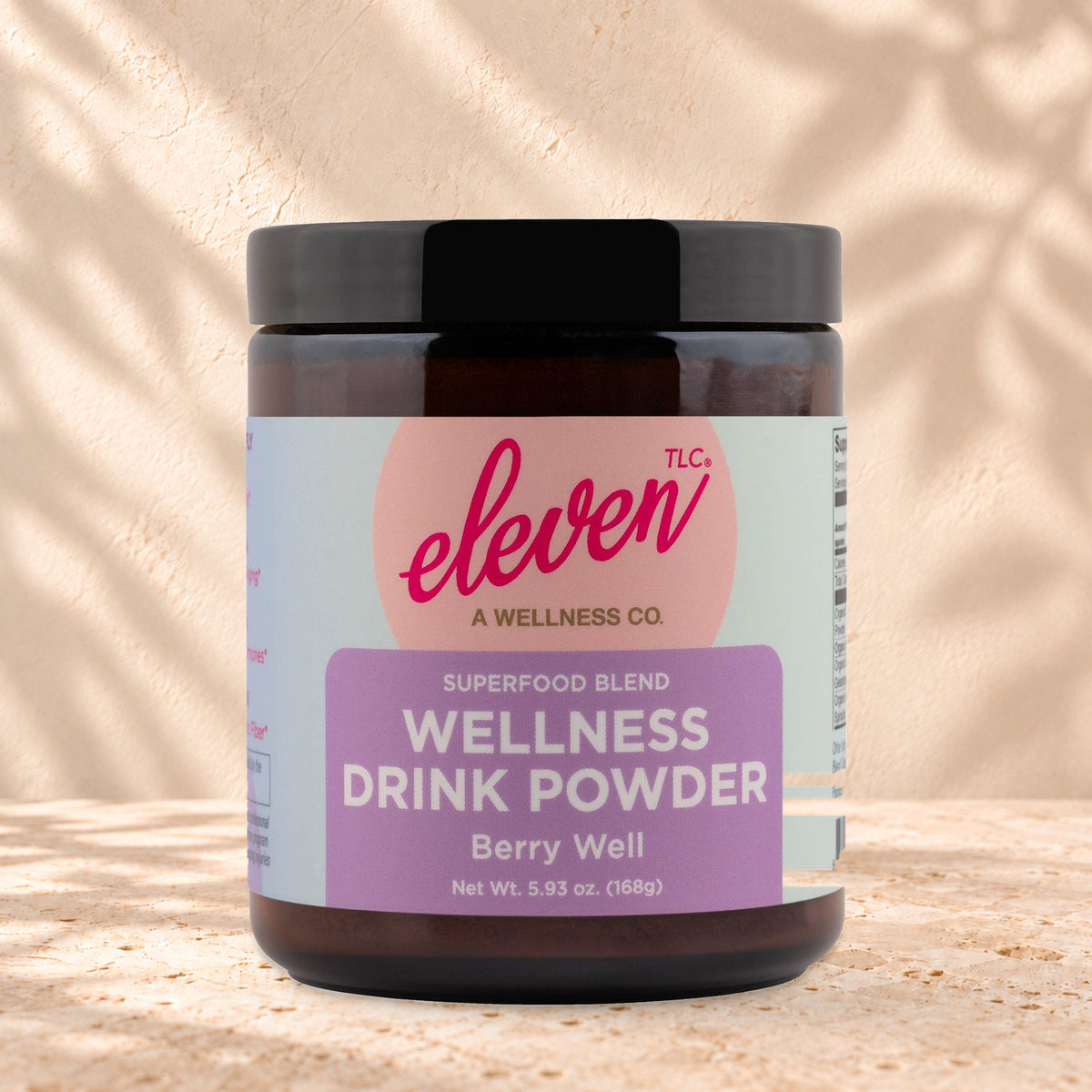 Eleven TLC Berry Well Superfood Latte Mix 168g amber glass jar with a black lid on travertine stone against a warm cream plaster wall, with a soft shadow to the right.
