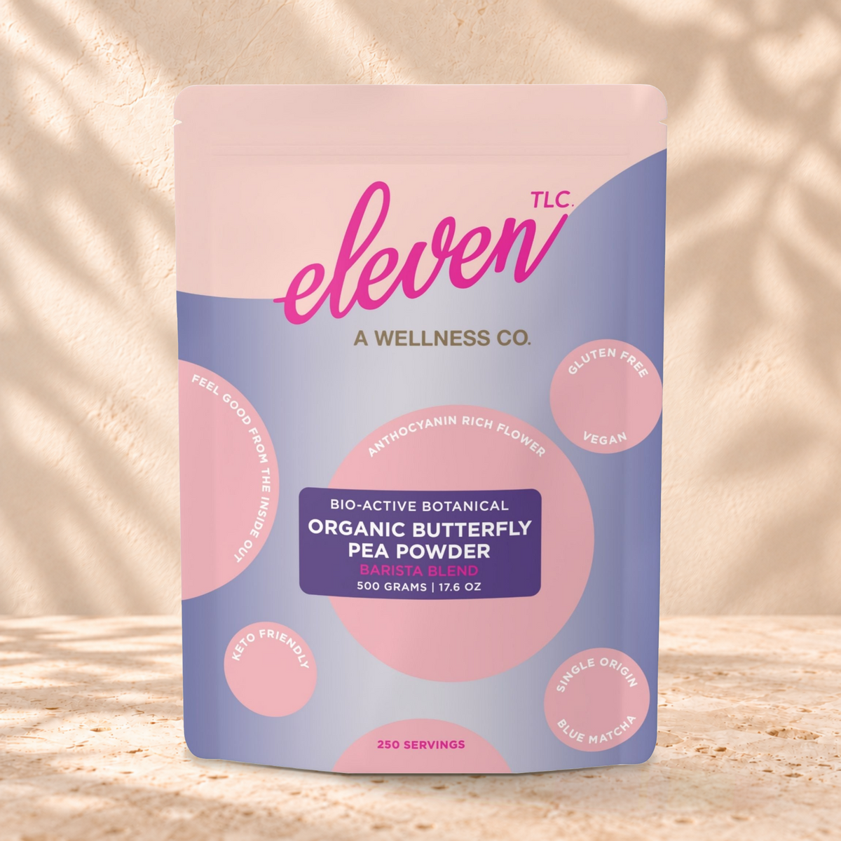 Eleven TLC Blue Matcha Butterfly Pea Powder 500g lavender and pink pouch standing on a travertine ledge against warm plaster, with a contact shadow beneath the base and a soft shadow to the right.