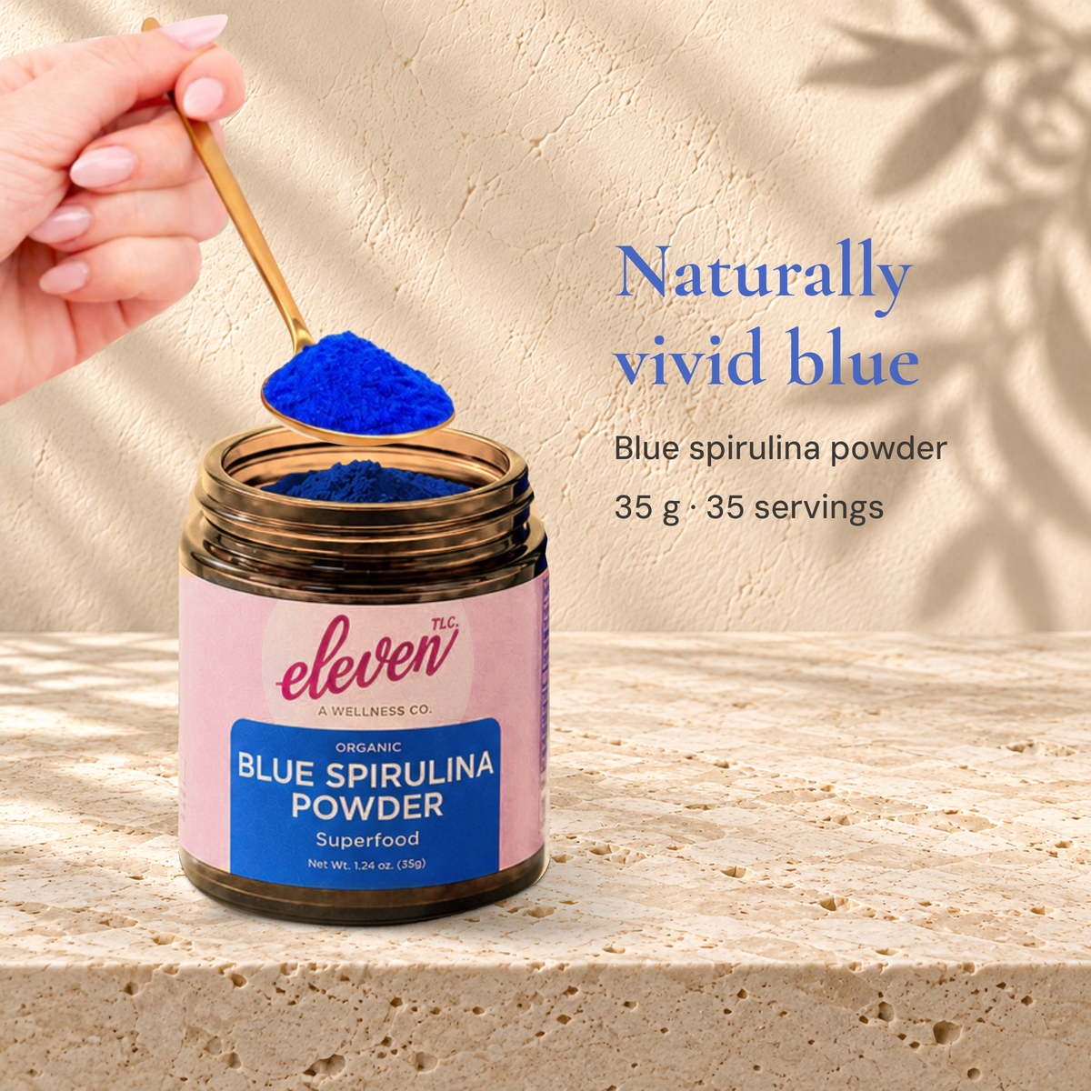 Open Eleven TLC Blue Spirulina Powder 35g jar beneath a hand holding a gold spoon of blue powder, on travertine.