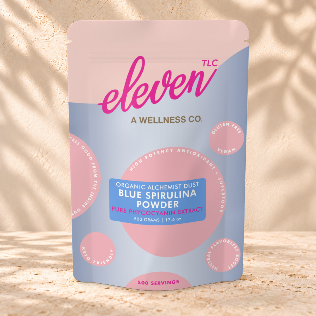 Front view of an Eleven TLC Blue Spirulina Powder 500g pouch on warm travertine against a cream plaster wall with botanical shadows.