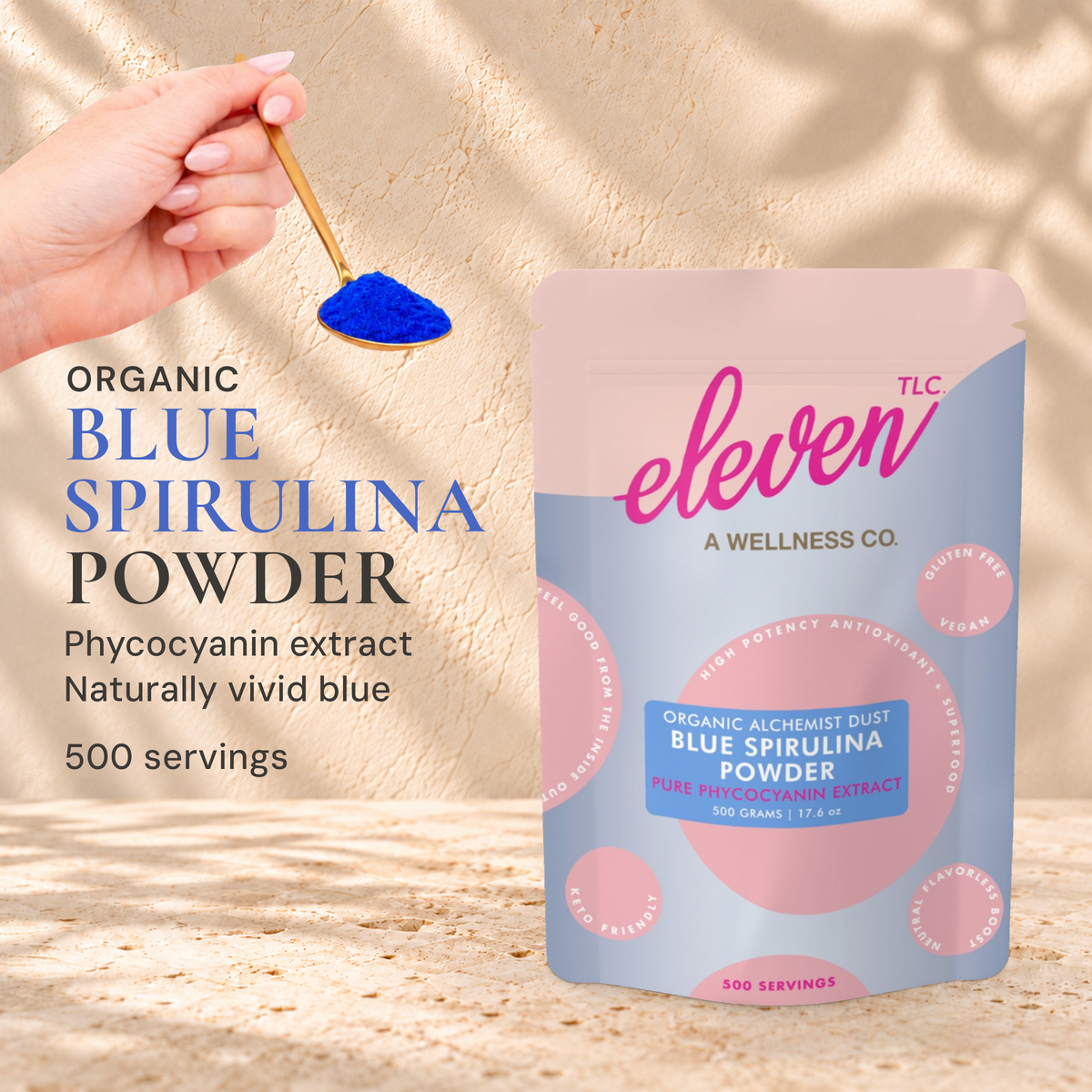 Eleven TLC Blue Spirulina Powder 500g pouch beside a hand holding a gold spoon of blue powder on warm travertine.