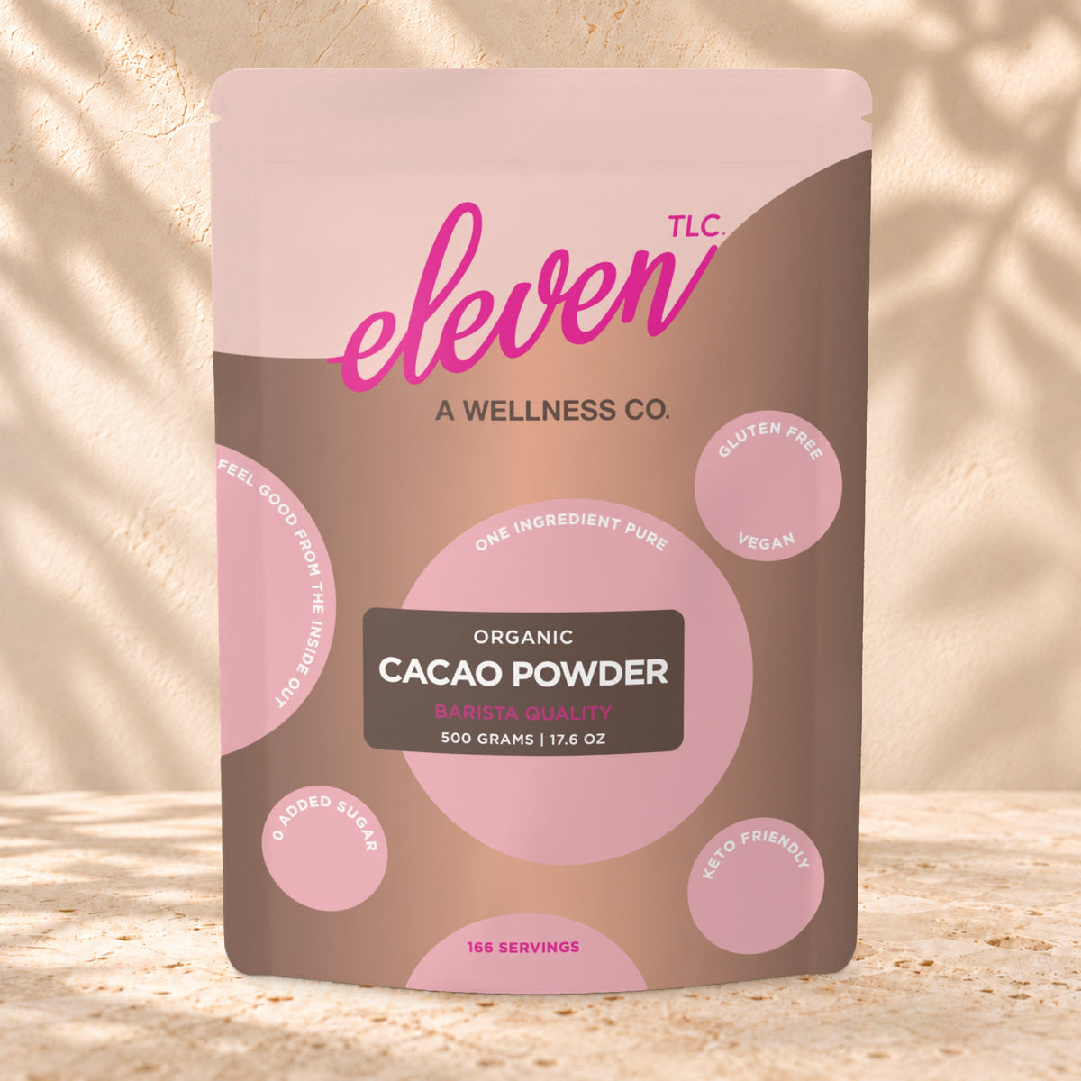 Front view of an Eleven TLC Cacao Powder 500g pouch on warm travertine against a cream plaster wall with botanical shadows.
