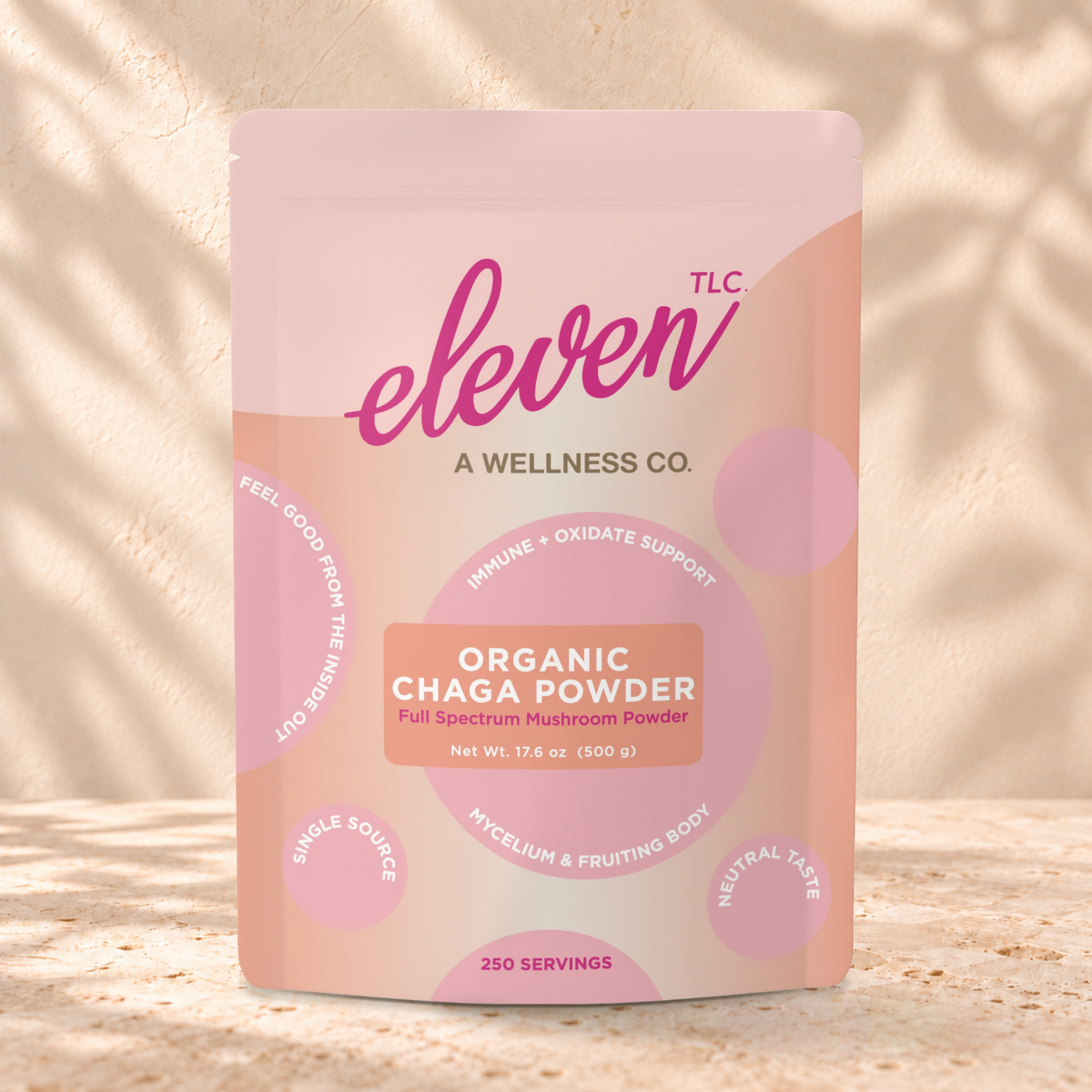 Eleven TLC Chaga 500g coral and pink pouch standing on a travertine ledge against warm plaster, with a contact shadow beneath the base and a soft shadow to the right.