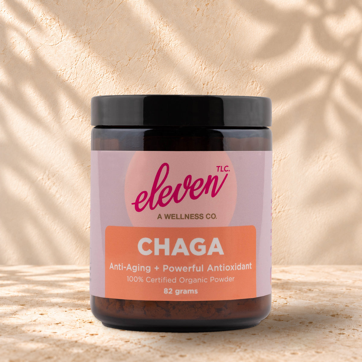 Front view of an Eleven TLC Chaga 82g amber jar with a black lid on warm travertine against a cream plaster wall, with a soft shadow extending right.