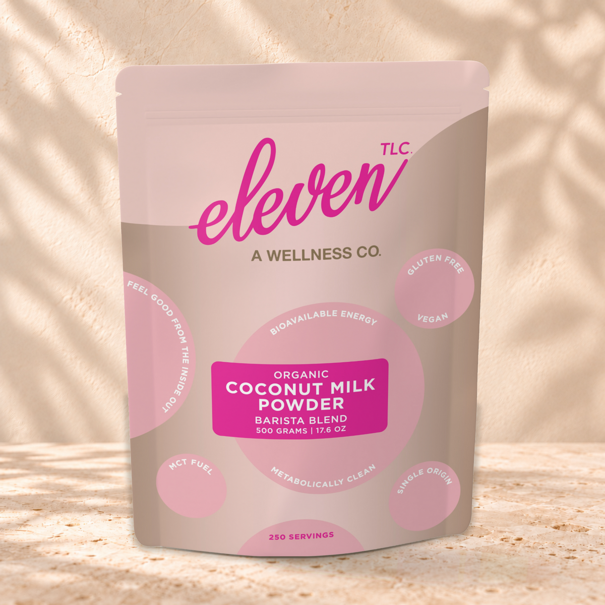 Eleven TLC Coconut Milk Powder 500g tan and pink pouch standing on a travertine ledge against warm plaster, with a contact shadow beneath the base and a soft shadow to the right.