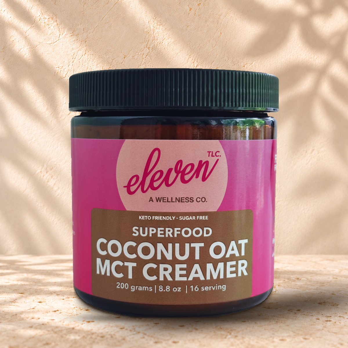 Front view of an Eleven TLC Coconut Oat MCT Creamer 200g amber jar with a black ribbed lid on warm travertine against a cream plaster wall, with natural contact shadows beneath the base.