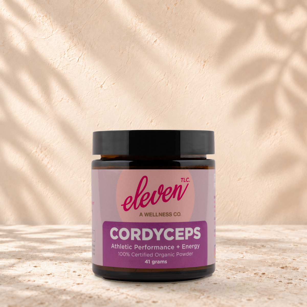 Eleven TLC Cordyceps 41 gram mushroom powder jar with a black lid standing on travertine against a warm plaster backdrop.