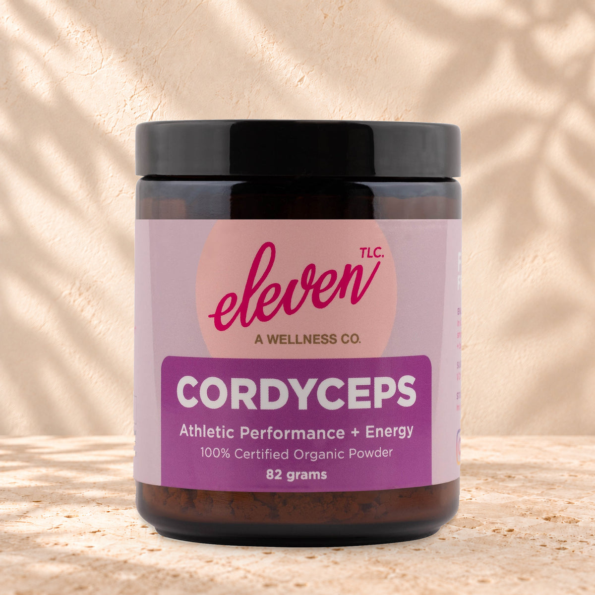 Eleven TLC Organic Cordyceps Mushroom Powder 82g amber glass jar with a black lid on travertine stone against a warm cream plaster wall, with a soft shadow to the right.