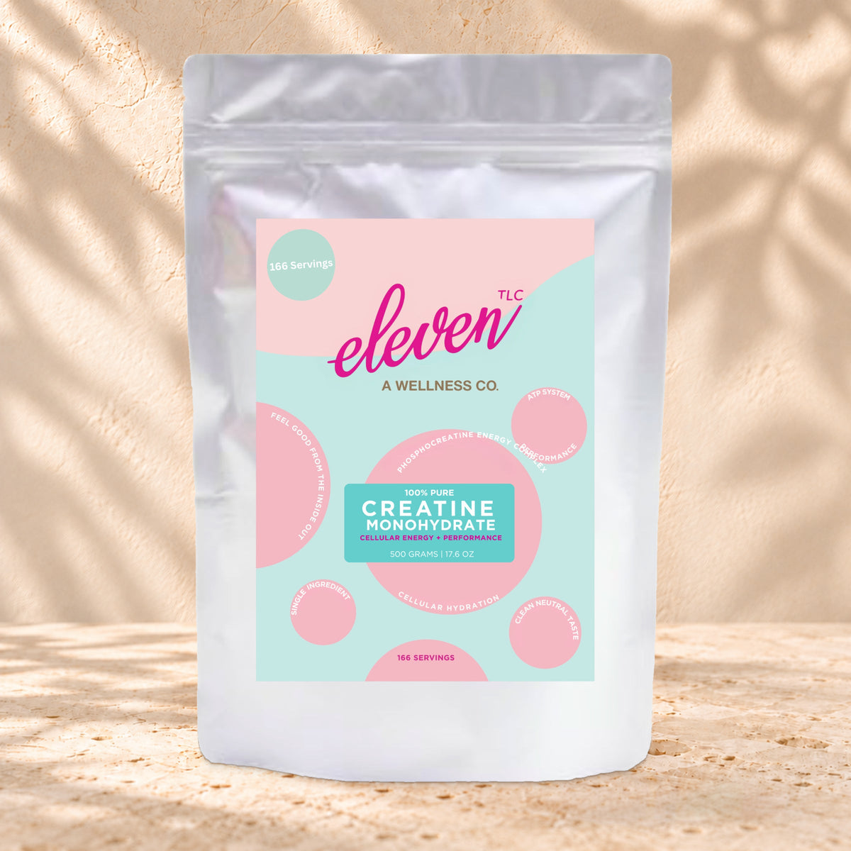 Front view of an Eleven TLC Creatine Monohydrate 500g silver pouch with a pink and aqua label on warm travertine against a cream plaster wall.