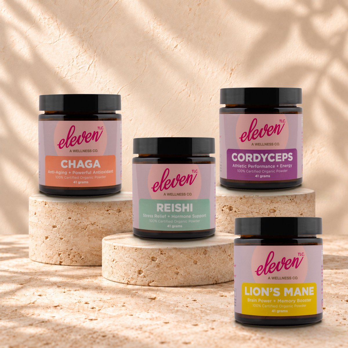 Four Eleven TLC organic 41 g mushroom powder jars—Chaga, Reishi, Cordyceps, and Lion's Mane—displayed on travertine risers against a warm plaster backdrop.