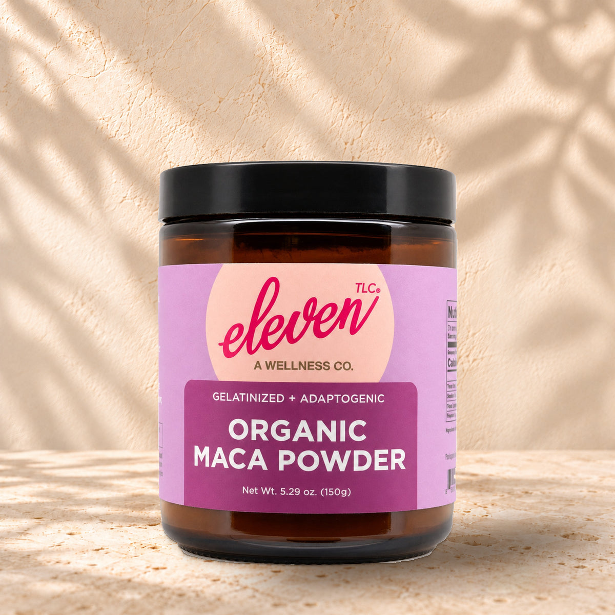 Front view of an Eleven TLC Maca 150g amber jar with a black lid on warm travertine against a cream plaster wall, with a soft shadow extending right.