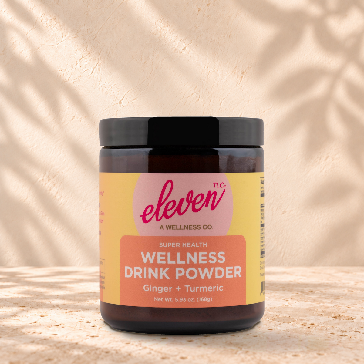 Eleven TLC Organic Ginger Turmeric Golden Latte Mix | Digestive Comfort, 168g jar in an amber glass jar with a black lid on a travertine ledge against warm plaster, with a slightly stronger contact shadow beneath the base and a soft shadow to the right.