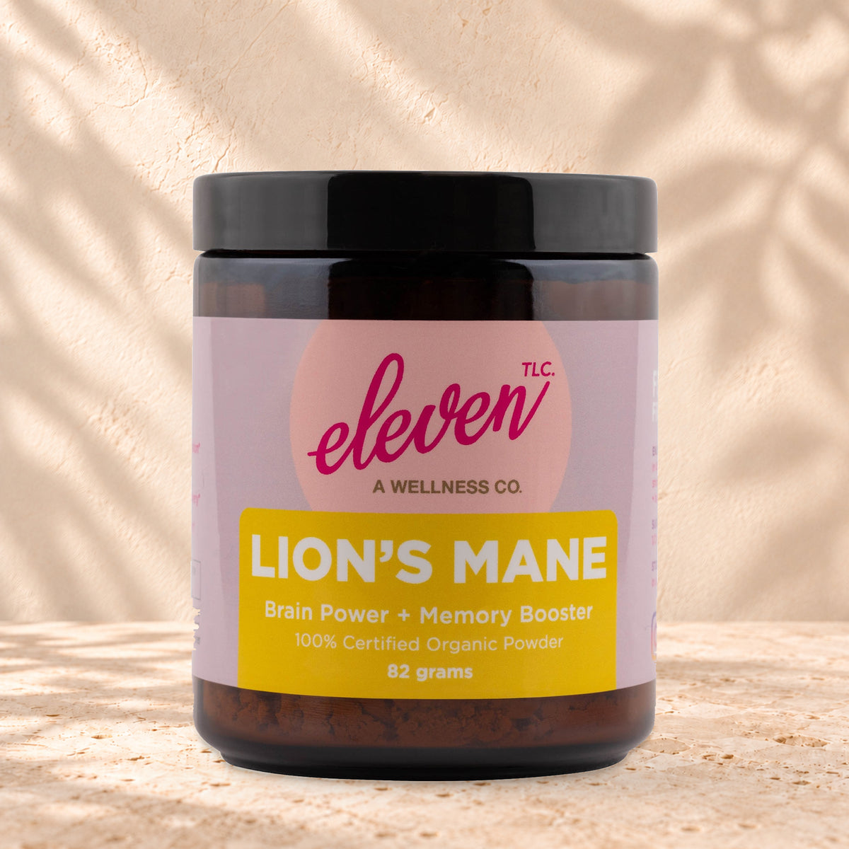Eleven TLC Organic Lion's Mane Mushroom Powder 82g amber glass jar with a black lid on travertine stone against a warm cream plaster wall, with a soft shadow to the right.