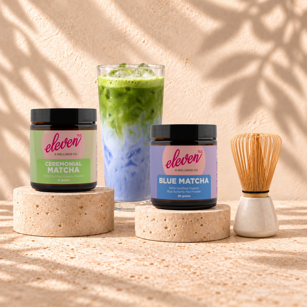 Eleven TLC Ceremonial Matcha 41g and Blue Matcha 36g jars with an iced layered matcha drink and bamboo whisk on travertine risers.