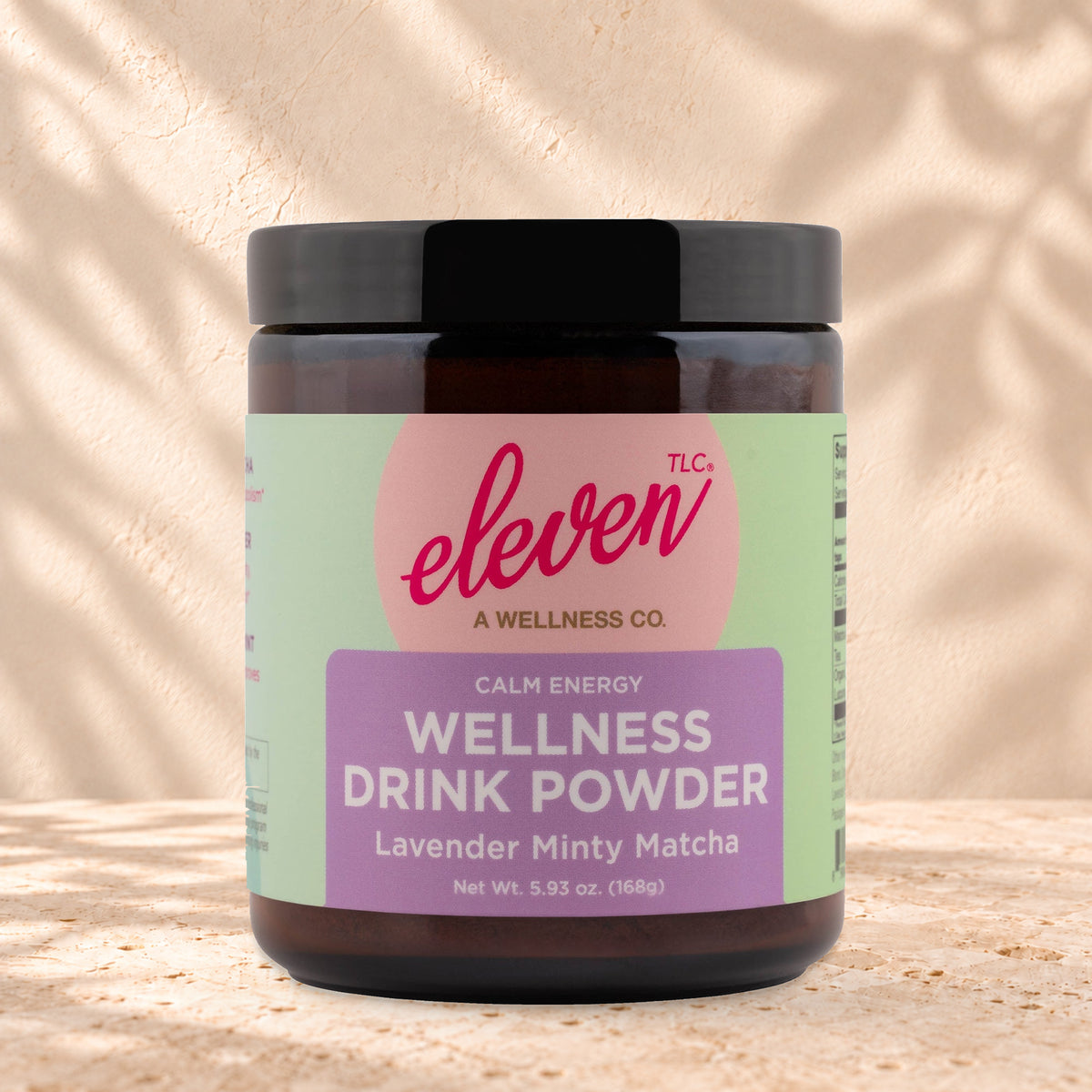 Eleven TLC Minty Lavender Matcha 168g amber glass jar with a black lid on travertine stone against a warm cream plaster wall, with a soft shadow to the right.