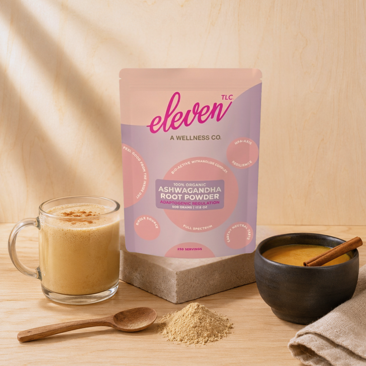 Eleven TLC organic ashwagandha root powder 500 g pouch on a stone block beside a glass mug of frothy latte, a wooden spoon of beige ashwagandha powder and a bowl of golden milk with cinnamon in warm window light.