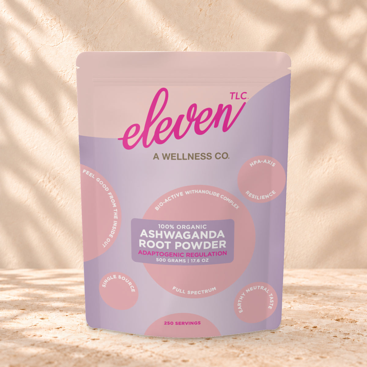 Eleven TLC Organic Ashwagandha Root Powder 500g stand-up pouch on travertine stone against a warm cream plaster wall, lit from the left with a soft shadow to the right.