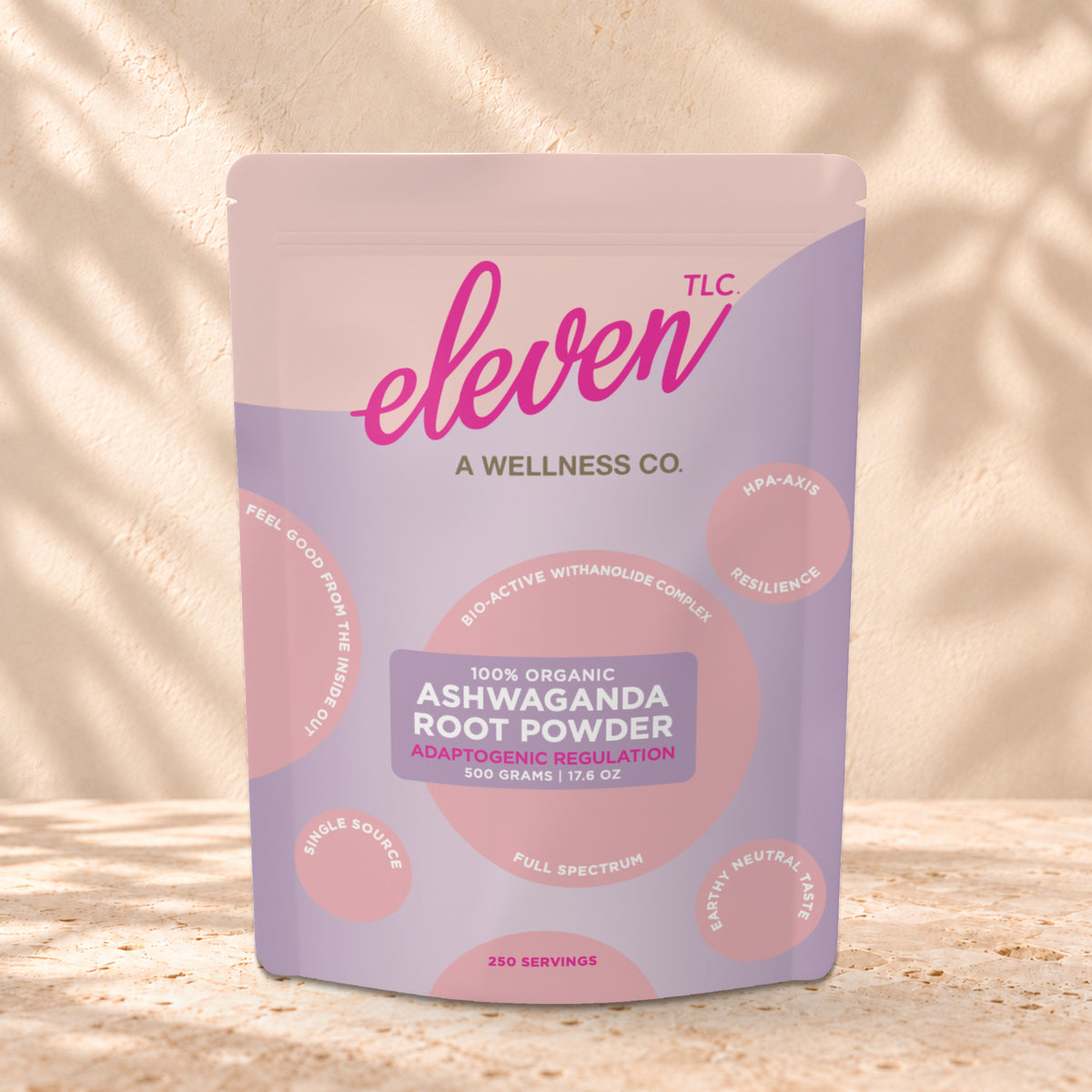 Eleven TLC Organic Ashwagandha Root Powder 500g stand-up pouch on travertine stone against a warm cream plaster wall, lit from the left with a soft shadow to the right.