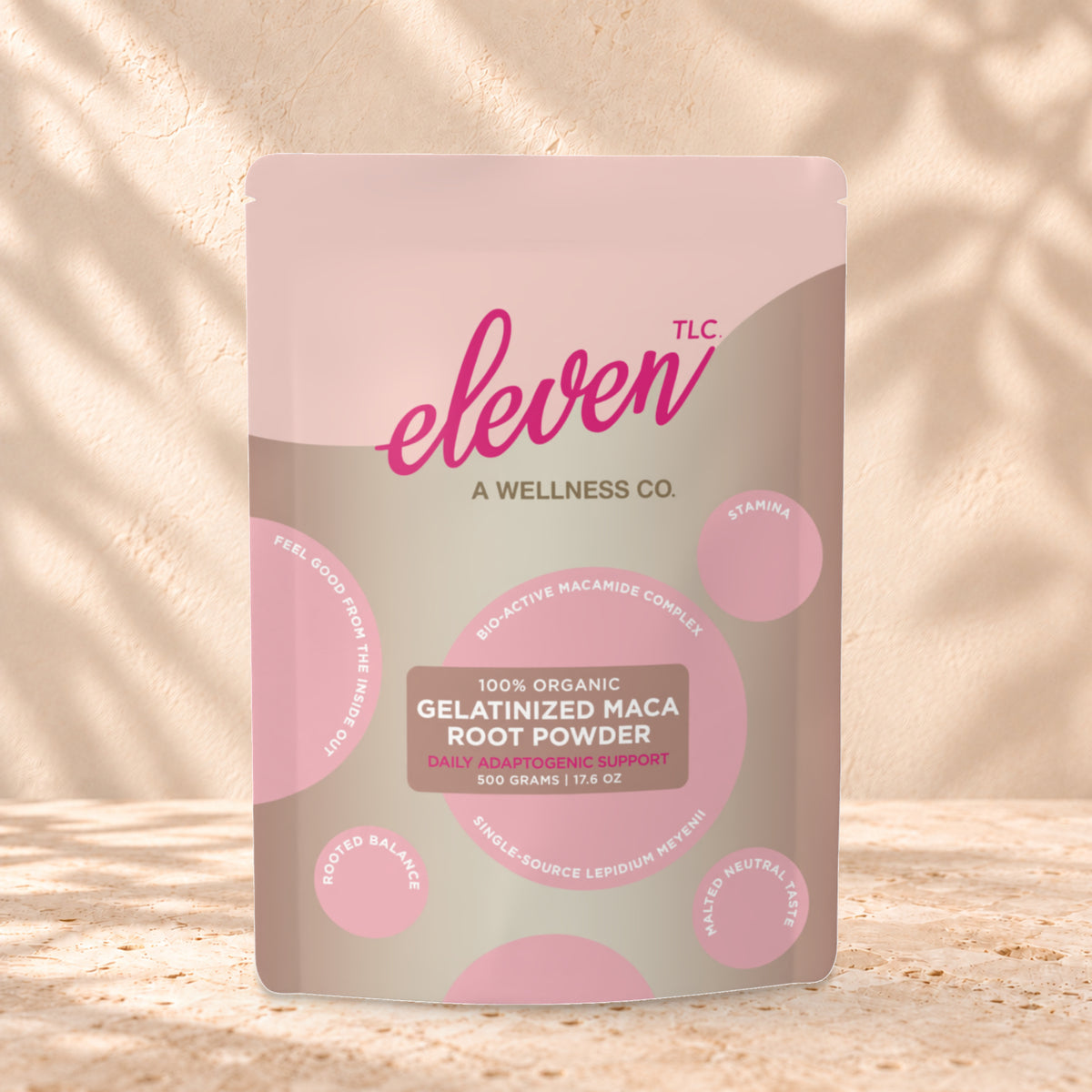 Eleven TLC Organic Gelatinized Maca Root Powder 500g stand-up pouch on travertine stone against a warm cream plaster wall, lit from the left with a soft shadow to the right.