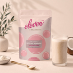 Eleven TLC organic MCT oil powder 500g pouch beside a glass of creamy milk, a bowl and gold spoon of white powder, coconut halves and an olive branch in warm light.