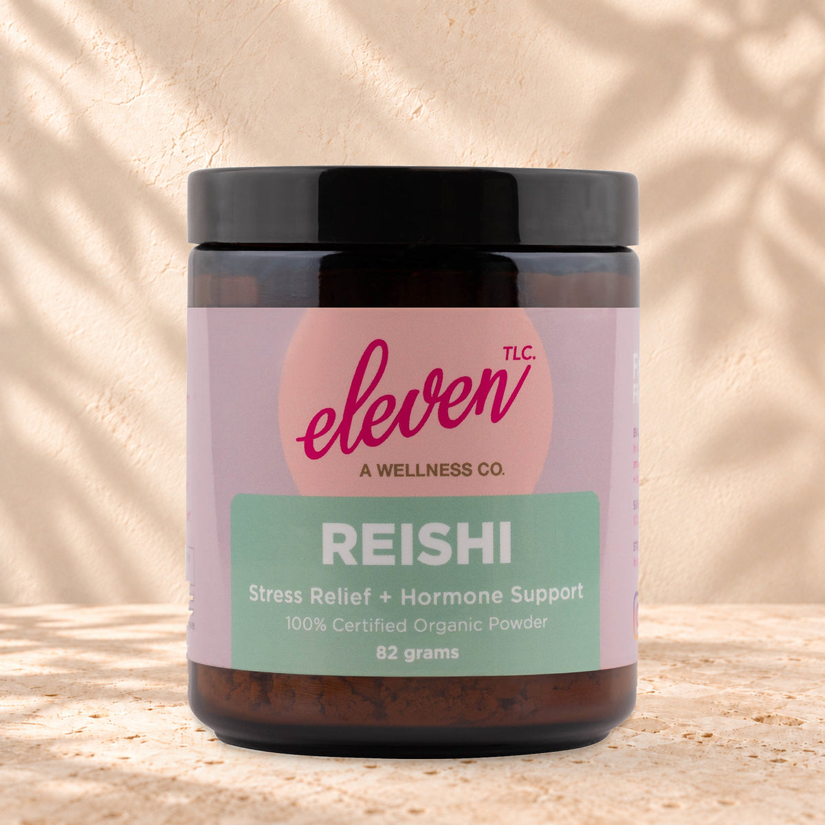 Eleven TLC Organic Reishi Mushroom Powder 82g amber glass jar with a black lid on travertine stone against a warm cream plaster wall, with a soft shadow to the right.