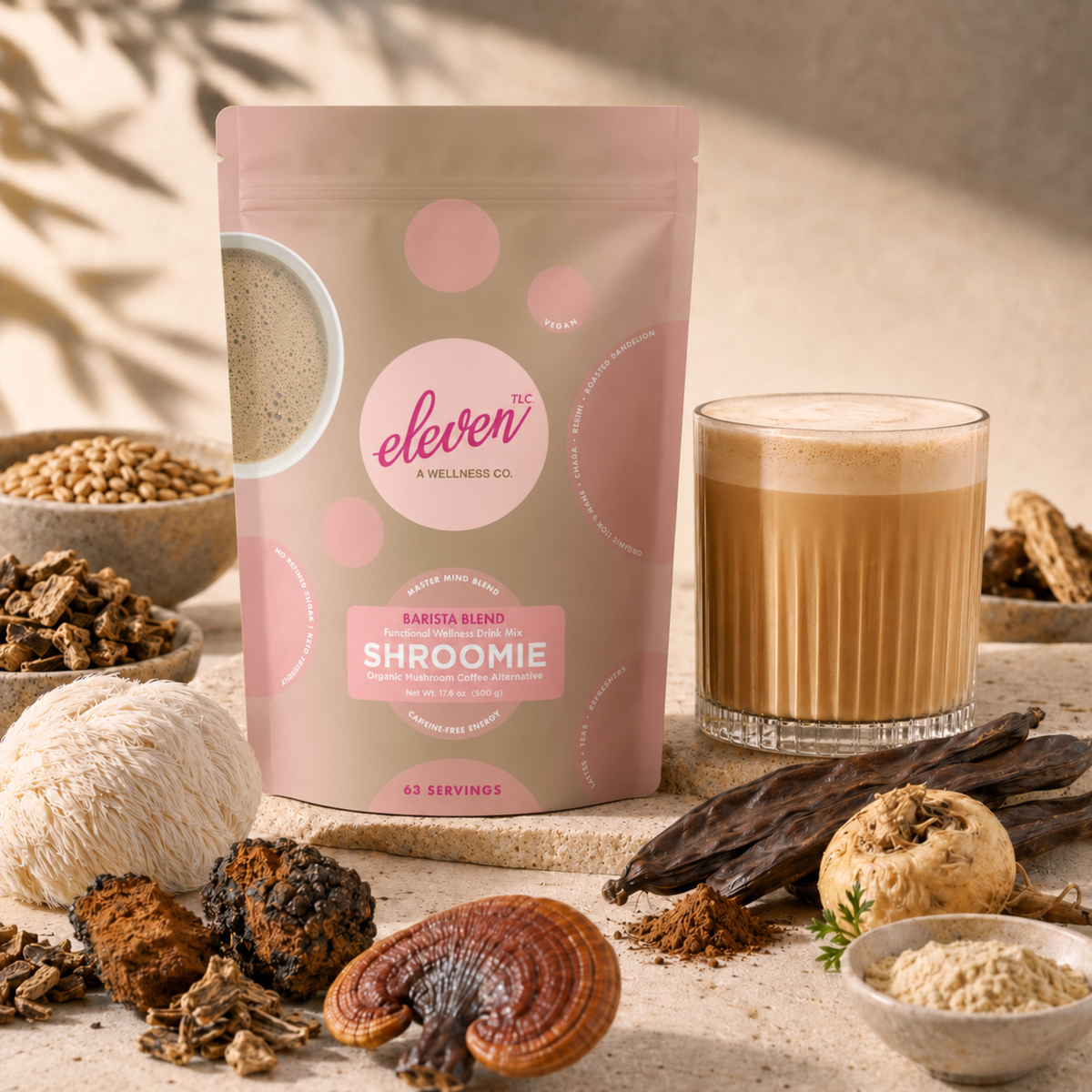 Eleven TLC Shroomie Barista Blend 500g pouch beside a latte in a ribbed glass on travertine, surrounded by roasted barley, chicory root, lion’s mane, chaga, reishi, carob pods and maca root