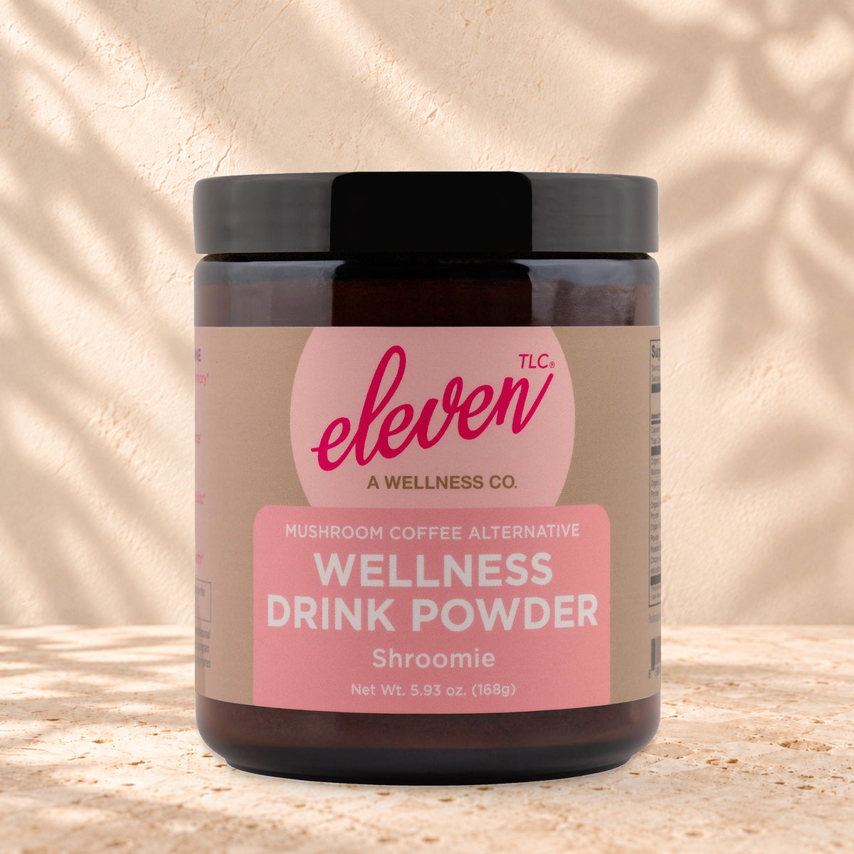 Eleven TLC Shroomie 8-Adaptogen Blend 168g amber glass jar with a black lid on travertine stone against a warm cream plaster wall, with a soft shadow to the right.